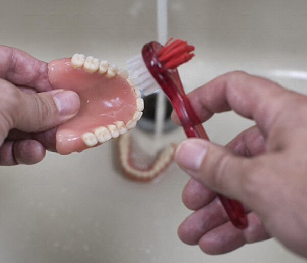 Hands using brush to carefully clean dentures over sink